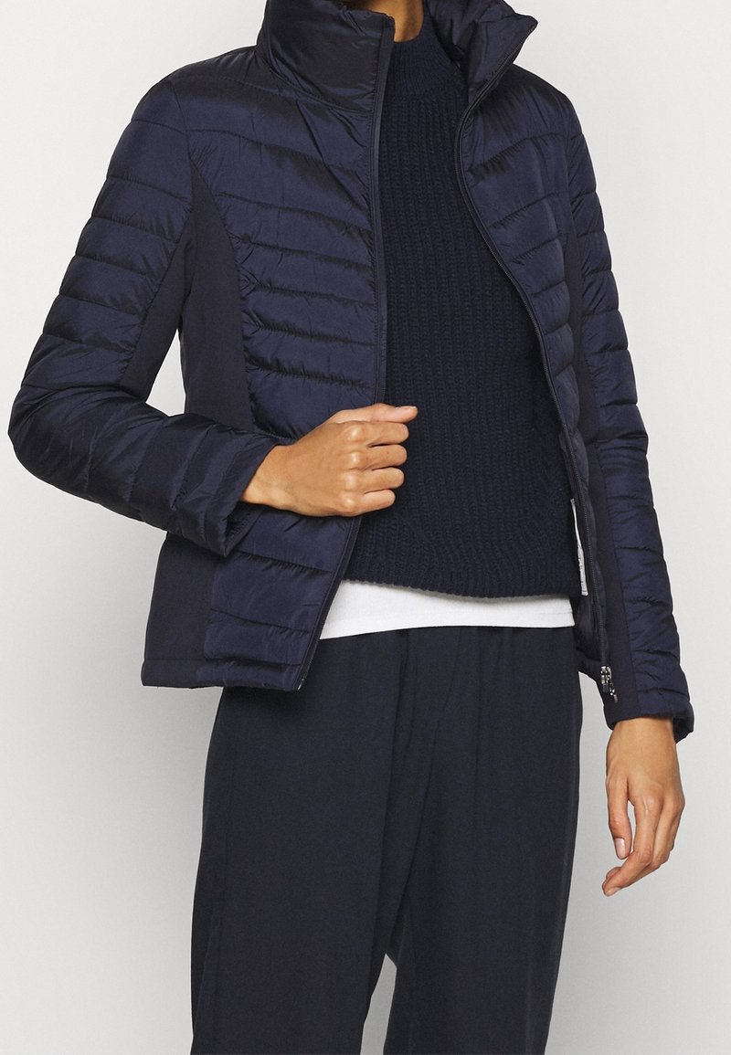 Giacca piumino blu navy con colletto alto, caratterizzata da una texture trapuntata e chiusura con zip. Indossata sopra un maglione di lana scuro.
