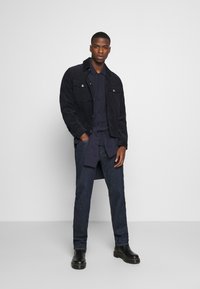 Chaqueta de pana azul oscuro con bolsillos en el pecho con botón, superpuesta a una camisa marina, combinada con jeans oscuros y botas negras de tobillo.