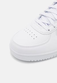 Valkoinen nahkainen urheilukenkä pyöreällä kärjellä, tekstuurilla varustetulla sivuosalla ja litteillä valkoisilla nauhoilla, joissa on pieniä ilmanvaihtoaukkoja kärjen kohdalla.