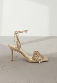 Gouden hoge hak sandaal met een vierkante tenen, franjes op de band en een strik rond de enkel. Glad uiterlijk, slanke stiletto hak.
