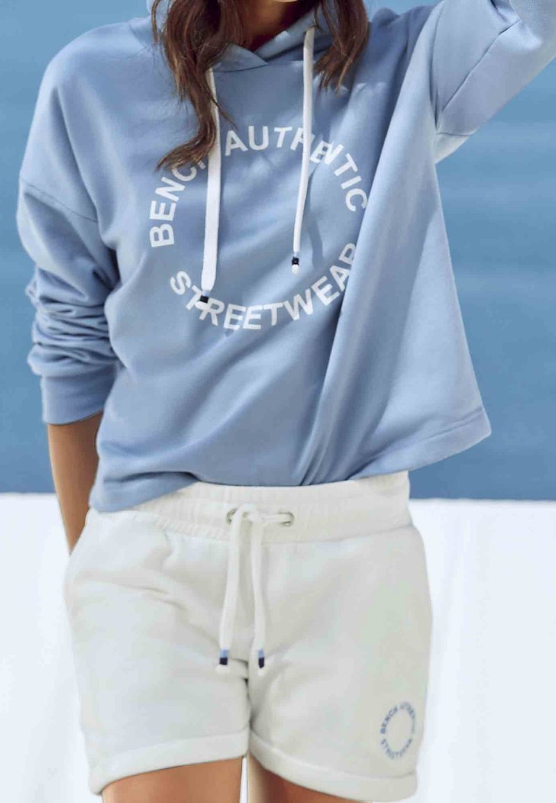 Personne portant un sweat à capuche bleu clair avec des cordons blancs et un short blanc avec un cordon, posant devant un fond bleu et blanc.