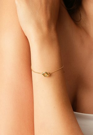 Bracelet délicat en chaîne d'or avec un petit pendentif en forme de nœud entrelacé sur le poignet d'une personne, sur une peau nue et un tissu blanc.