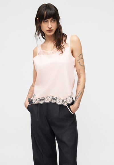 Γυναίκα με μακριά σκούρα μαλλιά και τατουάζ φοράει ένα ανοιχτό ροζ καμιζόλ με δαντέλα και άνετο σκούρο γκρι παντελόνι, με τα χέρια στις τσέπες.