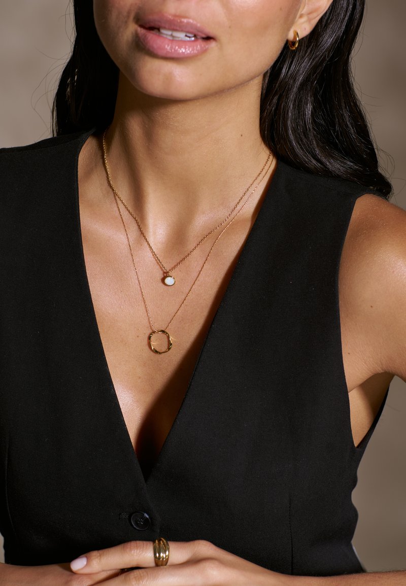 Colliers en or avec des pendentifs circulaires et en pierre superposés sur un haut noir en v. Le mannequin porte une bague en or et des boucles d'oreilles créoles. Texture de peau lisse.