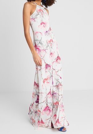 Kobieta ubrana w białą sukienkę maxi na szyję z różowo-fioletowym kwiatowym nadrukiem, w parze z niebieskimi sandałkami na otwartym palcu, stojąca na tle jednolitej szarej ściany.