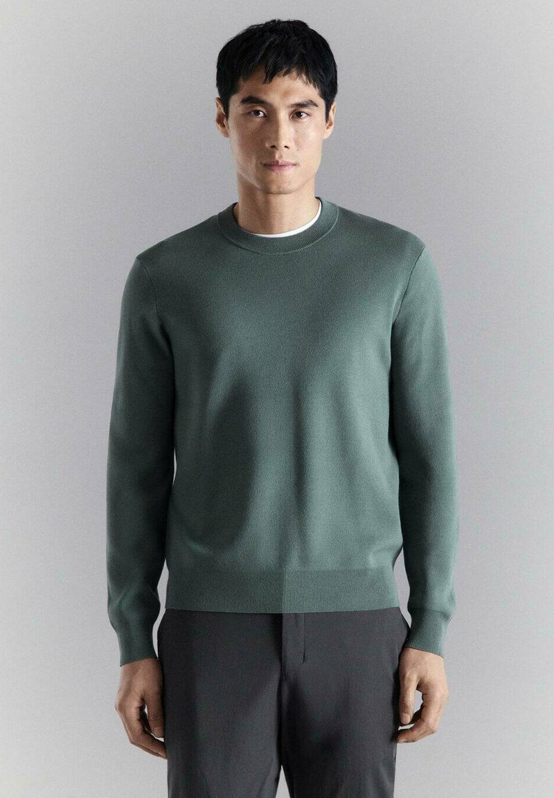 Man die een groene trui met ronde hals en donkere broek draagt, staand tegen een effen lichtgrijze achtergrond, vooruitkijkend met een neutrale uitdrukking.