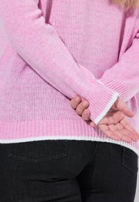Roze gebreide trui met geribde manchetten en witte afwerking, met een zachte, textuurrijke afwerking. Draag met zwarte spijkerbroek, met een achteraanzicht.