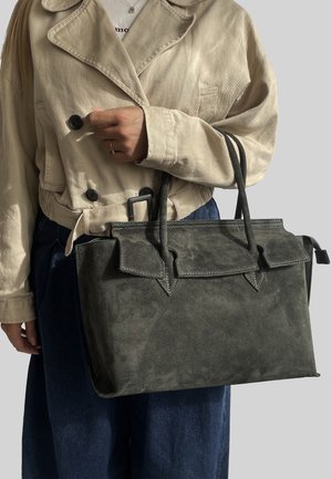 Borsa a tracolla in suede verde scuro con design strutturato, dettagli con pattina e due manici superiori, tenuta da una persona che indossa una giacca di colore chiaro.