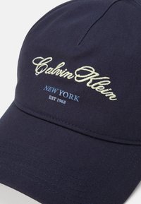 Gorra de béisbol azul marino con visera curvada. Presenta bordados de "Calvin Klein", "NUEVA YORK" y "EST 1968" en texto crema y azul. Tejido texturizado.