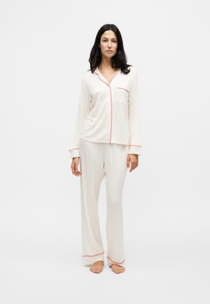Femme aux cheveux foncés portant un pyjama blanc à manches longues avec un liseré rouge, debout pieds nus devant un fond blanc uni.
