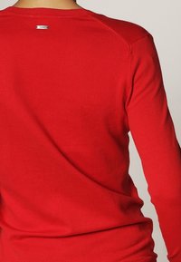 Pull rouge à manches longues en tissu tricoté lisse. Il présente un col rond et une étiquette logo discrète à l'arrière près du col.