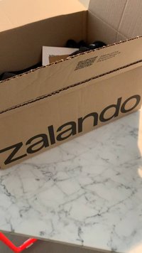 Kartoninė dėžė su žodžiu "zalando" parašytu ryškiomis juodomis raidėmis. Viduje matomi dalinai matomi daiktai ir QR kodas ant vienos pusės. Padėta ant marmurinio stalo.