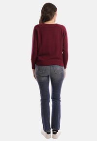 Donna in piedi di spalle, indossa un maglione bordeaux a maniche lunghe, jeans blu sbiaditi e sneakers bianche con calze nere.