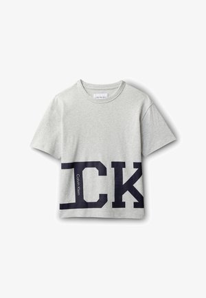 T-shirt a maniche corte grigio chiaro con grandi lettere nere "CK" e piccolo testo verticale "Calvin Klein" sulla parte inferiore frontale.