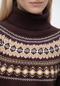 Gros plan d'une personne portant un pull à col roulé marron foncé avec des motifs géométriques crème, rose et moutarde autour de la poitrine.