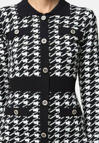 Cardigan à motif pied-de-poule noir et blanc avec col noir, ceinture noire et boutons décoratifs en argent sur le devant et les poches.