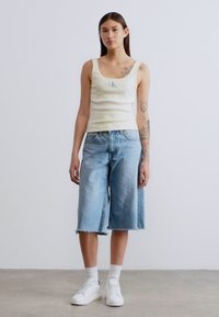 Ανοιχτόκκινο κίτρινο τοπάκι με μικρό λογότυπο, συνδυασμένο με ανοιχτό μπλε τζιν κολούς, λευκά αθλητικά παπούτσια και λευκές κάλτσες. Απλός, καθημερινός σχεδιασμός.