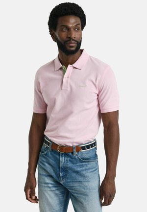 Light pink polo shirt with short sleeves and a green collar accent. Embroidered logo in light green. Worn with blue jeans and a brown belt.