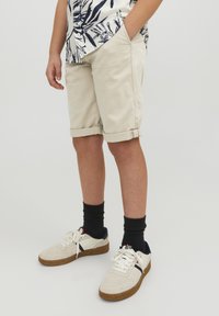 Pantaloni corti beige in cotone con risvolti, abbinati a sneakers bianche e calzini neri. La camicia presenta un motivo a foglie nere su sfondo bianco.