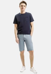 Granatowa koszulka z krótkim rękawem z kieszonką, w zestawieniu z jasnoniebieskimi szortami i białymi trampkami. Gładki materiał; luźny krój; swobodny design.