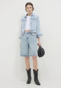 Giacca di jeans azzurra chiara e pantaloncini, top corto bianco, borsa nera, stivali cowboy neri al ginocchio con texture a motivo.