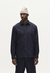 Donkerblauwe, geknoopte blouse van textuurstof met lange mouwen en een kraag, gecombineerd met bijpassende broek. Draagt een beige gebreide beanie.