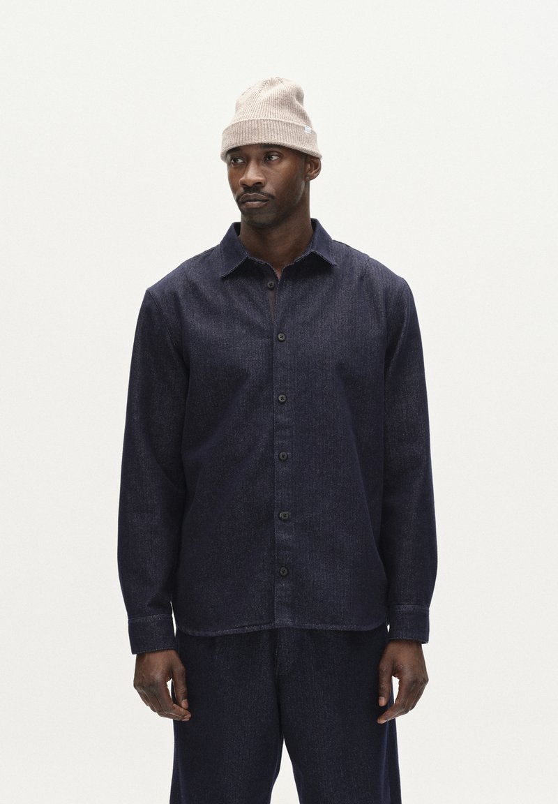 Donkerblauwe, geknoopte blouse van textuurstof met lange mouwen en een kraag, gecombineerd met bijpassende broek. Draagt een beige gebreide beanie.