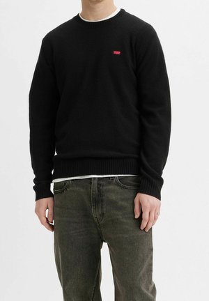 Man draagt een zwart trui met ronde hals en een klein rood logo op de borst, en vervaagde donkergrijze jeans, staand tegen een witte achtergrond.