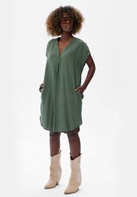 Zielona sukienka z krótkim rękawem i dekoltem w serek, z bokami kieszeniami oraz luźnym krojem, zestawiona z jasnobrązowymi botkami za kostkę.