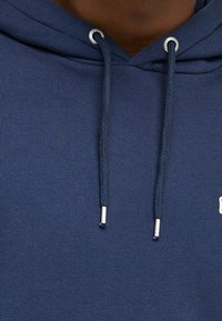 Sudadera con capucha de color marino hecha de tela suave, con ojales plateados y cordones redondeados con puntas metálicas. Logo destacado en la parte delantera.