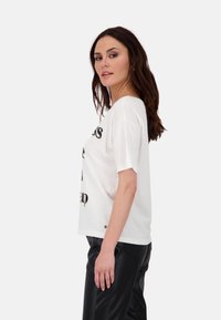 Weißes T-Shirt mit kurzen Ärmeln und grafischem Text, lockere Passform, aus Baumwollmaterial, getragen mit schwarzen Kunstlederhosen, Seitenansicht.