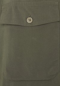 Olivgrüner Stoff mit einer Fronttasche, die über einen runden Knopfverschluss verfügt. Strukturiert mit sichtbaren Nahtdetails.