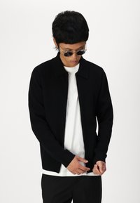 Svart zipped cardigan med krage, laget av teksturert stoff. Bæres over en hvit t-skjorte, med lange ermer og en avslappet passform.