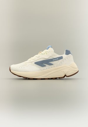 Weißer Sneaker mit blauem Akzent, klobiger beiger Sohle und "SHADOW"-Branding an der Seite, im Profil vor schlichtem Hintergrund gezeigt.