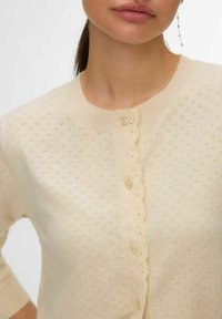 Cardigan crème avec patte de boutonnage festonnée, motif en tricot texturé et manches trois-quarts. Boutonné avec de petits boutons ronds.