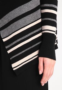 Maglione a righe nere e crema in maglia, con texture a contrasto, caratterizzato da ampie righe e polsini aderenti. La vista laterale mostra una mano che tiene il tessuto.