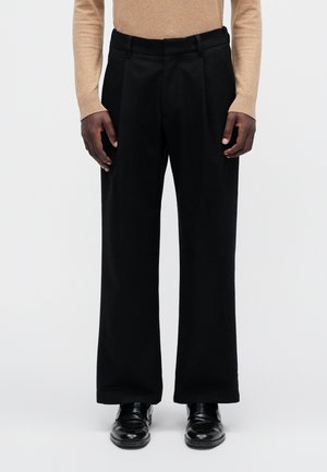 Mężczyzna ubrany w czarne spodnie o szerokich nogawkach, beżową bluzkę z długim rękawem oraz czarne skórzane eleganckie buty na tle jednolitego tła.