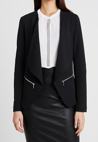 Svart blazer med draperad front, två dragkedjefickor och skräddarsydd passform. Bärs över en vit blus med en silveraccent. Texturerad svart kjol.