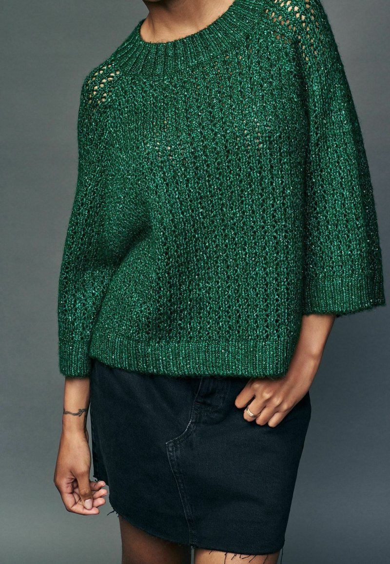 Pull en laine verte avec un tricot lâche et des poignets côtelés, assorti à une jupe en denim noire. La texture est douce et légèrement transparente.