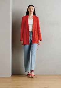 Blazer rosso, top bianco e jeans a gamba larga azzurri chiari. Il modello indossa sandali rossi e si trova davanti a una parete grigia liscia con pavimento in legno.