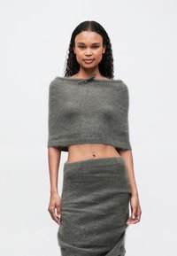 O femeie cu păr creț poartă un top scurt gri, pufoș, cu umerii la vedere și o fustă asortată, de culoare gri, cu talie înaltă, stând în fața unui fundal alb simplu.