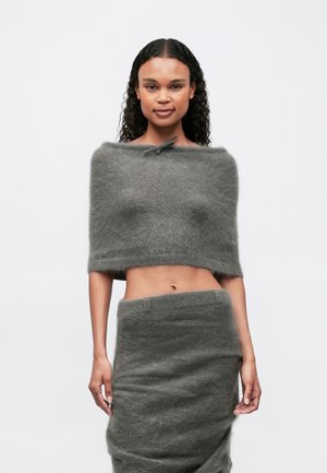 Kvinne med krøllete hår har på seg en grå, fluffy avskåret topp uten skuldre og matchende høy midje skjørt, står mot en enkel hvit bakgrunn.