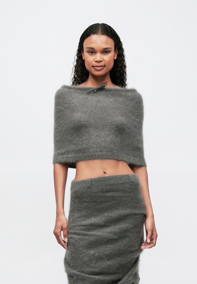 O femeie cu păr creț poartă un top scurt gri, pufoș, cu umerii la vedere și o fustă asortată, de culoare gri, cu talie înaltă, stând în fața unui fundal alb simplu.