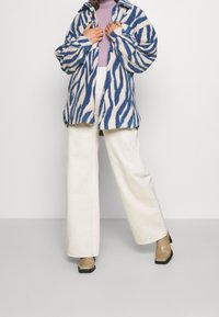 Niebiesko-biała bluza polarowa w wzór zebry o luźnym kroju, zapinana na guziki, z dużym kołnierzem, zestawiona z białymi spodniami o szerokich nogawkach.