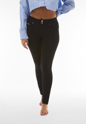 Kobieta w czarnych dopasowanych spodniach i krótkim jasnoniebieskim długim rękawem, stojąca boso na białym tle.