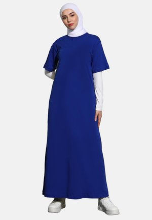 Modrá maxi šaty s krátkými rukávy, vyrobené z hladkého materiálu. Nosené s bílým hidžábem a kombinované s bílými teniskami. Jednoduchý design, bez vzorů.