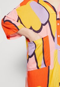 Kolorowa bawełniana koszula z dużymi abstrakcyjnymi wzorami kwiatowymi w odcieniach pomarańczowym, żółtym, różowym i fioletowym, z zapięciem na guziki z przodu i boczną kieszenią.