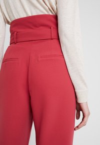 Červené kalhoty s vysokým pasem a širokým pasem, se dvěma zadními kapsami a hladkou texturou látky, nošeny s světlým béžovým topem.