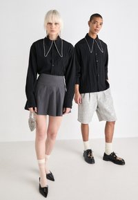 Camisa preta de botão com colarinho oversized e detalhe em corrente prateada, saia plissada cinza, calções cinzentos com lantejoulas e sapatos pretos.
