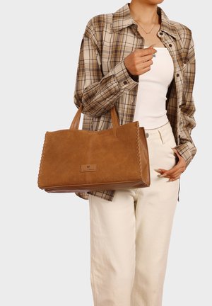 Persona che indossa una camicia a quadri beige e pantaloni color crema che porta una grande borsa tote marrone in camoscio con bordi cuciti.
